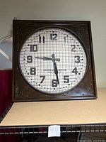 Front view of the square wall clock showing its white face and black numbers behind a grid pattern with American Clock Co. logo and model number LUG-44 on the face.