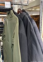 Side view of four hanging men's outerwear items including a textured green shirt with zippered collar, blue blazer, grey wool coat, and black wool coat.