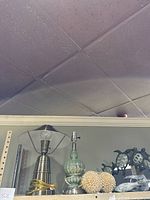 Photo showing green glass lamp base with metal fittings, small metal nautical anchor sculpture, metal sculpture of three sea turtles, and seashell balls in netting on shelf.