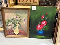 Two oil paintings side by side on the floor: one framed with white and purple flowers in a glass vase; the other unframed depicting bright pink roses in a blue vase on green background.