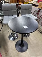 Two grey faux leather adjustable bar stools with padded seats and chrome bases positioned behind a small round black wood top table with a black pedestal base. The table shows minor scratches on the surface.