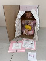 Porcelain doll in original box packing, wrapped partially in protective material, showing certificates of authenticity on table.