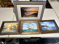 Three framed oil paintings of nautical themes: harbor with boats, sailing ship at sea, and lighthouse scene, each in wooden frames.