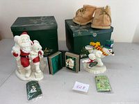 Multiple collectible figurines including Snowbabies and Santa, small framed ornaments, a pair of teddy bear boots in original boxes, and a small ornament pack arranged on white surface.