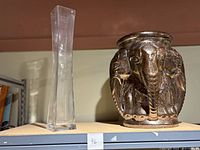 Two items on a shelf: a tall clear square glass vase and a ceramic pedestal with elephant heads painted in metallic black and gold.