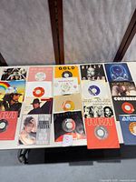 Top-down view of 20 vintage 45 RPM vinyl records from multiple artists in their original sleeves, arranged on a table. Sleeves and records show signs of age, wear and need cleaning.
