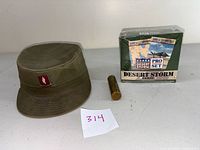 Photo of the vintage military hat with visible red Rhode Island National Guard insignia patch, beside a sealed box of Desert Storm collectible cards and a brass lighter case.