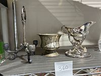 Three vintage silver-plated items on a metal shelf: a tri-candelabra, a metal champagne urn, and an ornate gravy boat on a warming stand.