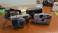 Two vintage cameras side by side on a table with carrying straps and a small black leather bag.
