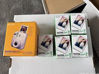 Photo showing Fujifilm Instax Mini 12 camera box in lilac purple along with five unopened boxes of Fujifilm Instax mini film.