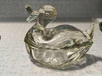 Clear depression glass duck figurine angled front view showing detailed head and body lid with visible chip on the base