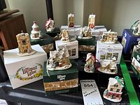 Twelve Lilliput Lane Christmas ornaments shown with original boxes, arranged on a black surface, displaying the detailed miniature houses and shops covered with snow and festive decorations.