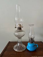 Both oil lamps shown side by side on wooden surface. Taller clear glass lamp on left, shorter blue ceramic base lamp on right.