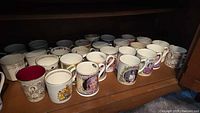Shelf displaying approximately 30-40 royal commemorative mugs with portraits and crests, varying sizes and styles