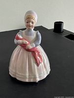 Front view of Royal Doulton The Rag Doll porcelain figurine showing young girl holding a rag doll wrapped in red cloth, wearing bonnet and pastel dress.