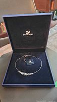 Swarovski necklace inside black box with Swarovski logo, showing front view of necklace with crystals aligned on wire and clasp visible.