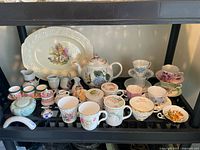 Full lot view showing various teacups with saucers, mugs with lids, teapot, and serving platter arranged on black shelves