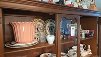 Wooden cabinet shelf with pink ceramic vase, decorative plates with vintage and royal designs, clear glass containers, and white cutout vase