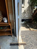 Wood push broom leaning vertically against a wall outdoors with some leaves near the bristles