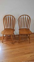 Front view of two wooden Windsor style chairs made of maple with spindle backs and contoured seats, placed on wooden floor against white wall.