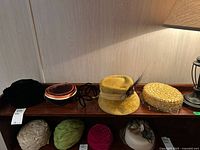Nine vintage ladies hats arranged on a wooden surface, showing variety in materials and colors: black velvet, yellow wool, brown fabric, green, cream, pink, and straw with netting.