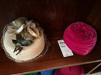 Two vintage hats photographed on wooden shelf: beige straw with flowers and veil, pink velvet rosette hat