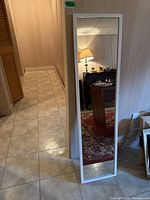 Mirror standing against a wall, showing full length and reflection of a room with carpet and furniture.