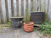 View of two large resin planters (dark color) and one smaller terra cotta planter with soil inside, situated outdoors against a wooden fence.