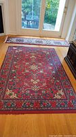 Full view of rectangular area rug on hardwood floor with geometric and floral border and red central field.