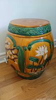 Full view of round ceramic side table/stool with orange body and raised green and white floral motifs, top is tan.