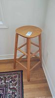 Wooden bar stool shown from a side view, standing on hardwood floor next to a wall and patterned floor rug.