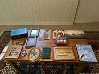Wide angle view of all items arranged on a wooden table including framed prints, plaques, decorative eggs in glass case, box and small trinkets.