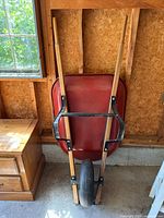 Wheelbarrow resting upright against a wooden wall, showing wooden handles, red metal body, and black metal supports on the wheel.