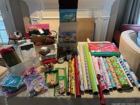 Wide view displaying numerous rolls of holiday and occasion-themed wrapping paper, gift bags, boxes, ribbons, and other supplies on a table including some partial rolls and packets.