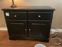 Front view of black wooden sideboard showing two drawers and two doors with silver-tone knobs and handles, sitting on hardwood floor.