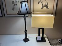 Photo showing two lamps on a white table against a wall with framed art. Left lamp is black metal with beige fabric shade. Right lamp is black bronze with rectangular beige fabric shade.