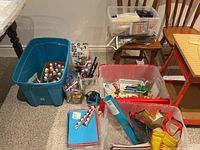 Overview of multiple bins and containers filled with assorted arts and crafts supplies including paint bottles, papers, brushes, glue, and assorted tools and embellishments lined up on floor and chairs.