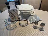 Full lot of assorted kitchen tools including glass measuring cup, grater, bowls, funnels, shakers, and peeler on table.