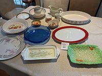 Overview of lot containing assorted serving platters, dishes, and pitchers displayed on a table showing mixed ceramic, porcelain, glass and stoneware items.