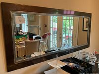 Full front view showing rectangular wood framed mirror with studded detail. Reflects room with dining and living areas.