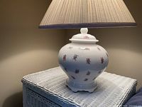 Full view of the vintage ceramic lamp with floral design and fabric shade showing the lamp lit on a white wicker table.