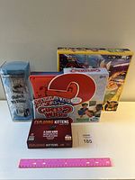 Front view of four games: Find It Skill Tester cylindrical game, Pokemon Battle Academy box and components, Guess Who? board game box, Exploding Kittens card game box and rulebook, ruler for scale.