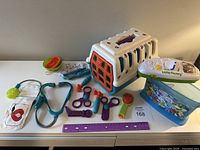 Wide shot of vet kit items including plastic pet carrier, veterinary tools, LeapFrog Letter Factory toy, and purple ruler laid out on white surface.