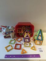 Photo of colorful magnetic geometric building tiles including squares and triangles, some assembled into a shape, with a red plastic storage bin and a ruler for scale.