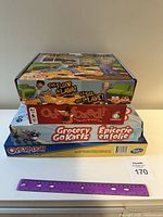 Stack of four boxed children's board games including The Floor is Lava, Outfoxed!, Grocery Go Karts, and Operation, with purple ruler for scale.