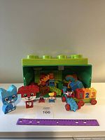 Overall photo showing the green Lego brick storage box, assorted Duplo blocks and characters like a blue cat and red dog, three small figures including a girl and boy, and a vehicle with 1-2-3 blocks.