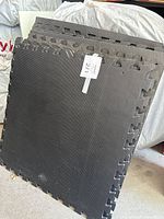 Photo 1 showing stack of large black rubber interlocking flooring mats leaning against a white plastic sheet and a bag.