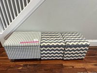 Three fabric storage cube ottomans side by side on a wooden floor against a white wall and railing.