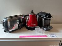 All three appliances (Oster toaster, De Longhi kettle, Nespresso machine) with two clear cups placed on white countertop with pink ruler for scale.