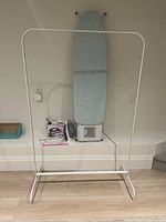 White IKEA hanging rack frame and ironing board with new blue cover set against a white wall and wooden floor